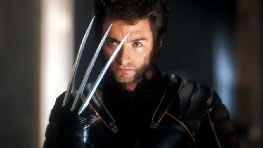 A man with sideburns and thick hair wears a black suit and raises his hand, displaying three long metal claws that extend from between his knuckles. He has a serious expression and is indoors.