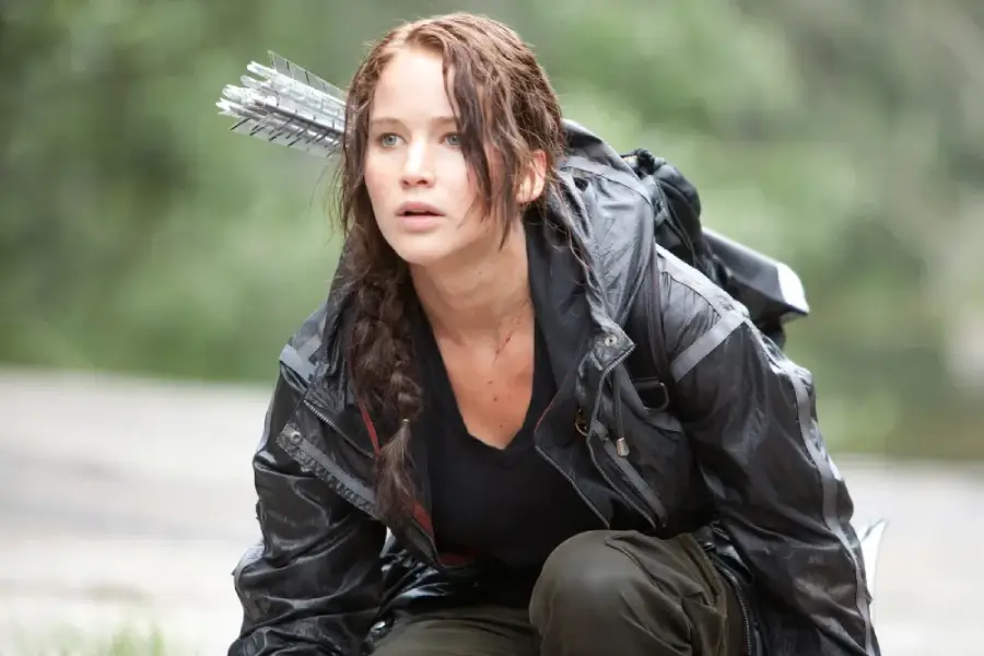 A young woman with a braided hairstyle crouches outdoors, wearing a black jacket and carrying a quiver of arrows on her back, looking alert and focused.