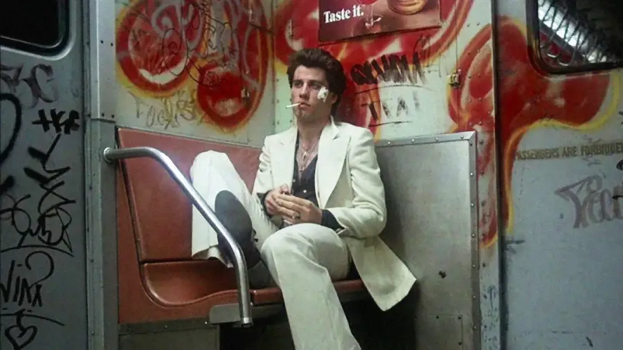 A man in a white suit with a bandage on his face sits alone on a graffiti-covered subway bench, holding something in his hands. The wall behind him is full of colorful spray paint and a torn advertisement.