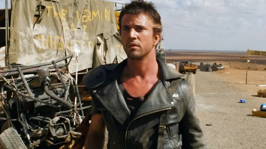 A man in a black leather jacket stands on a deserted road in a barren, post-apocalyptic landscape, with wrecked vehicles and a makeshift sign that reads "THE VERMIN HAVE INHERITED" in the background.
