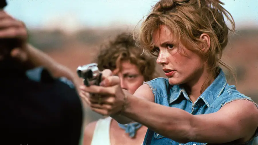 A woman in a denim sleeveless shirt points a handgun intently, with another woman standing behind her, both outdoors in a tense scene.