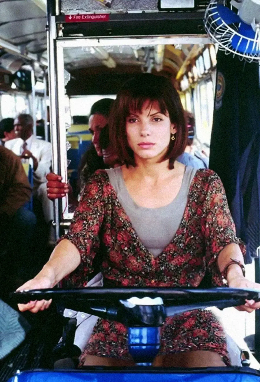 A woman with short brown hair, wearing a floral patterned top, sits at the wheel of a bus, focused and alert. Several passengers are seated behind her inside the bus.