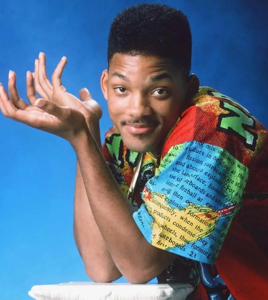 A young man with a high-top fade hairstyle wears a colorful, patterned shirt and leans on a pedestal, shrugging with an amused expression against a blue background.