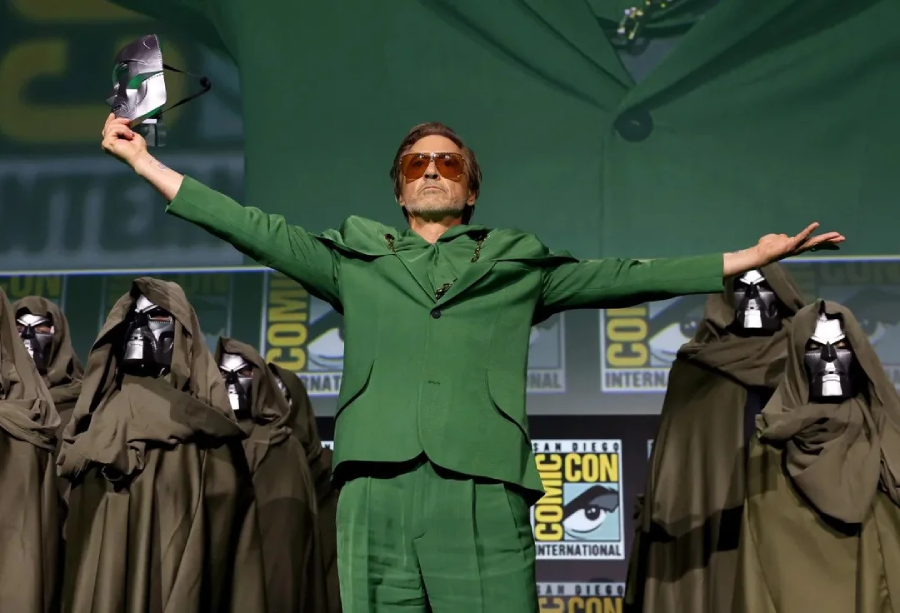 A man in a green suit stands center stage with arms outstretched, surrounded by people in dark cloaks and metal masks. He holds a mask in one hand. Comic-Con International logos are visible in the background.