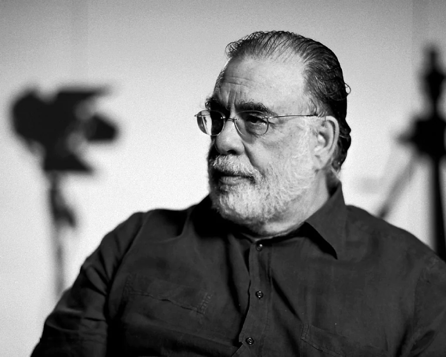 A man with glasses and a beard is sitting and looking to the side. He has slicked-back hair and is wearing a dark shirt. The background is out of focus with studio equipment visible in the shadows. The image is black and white.