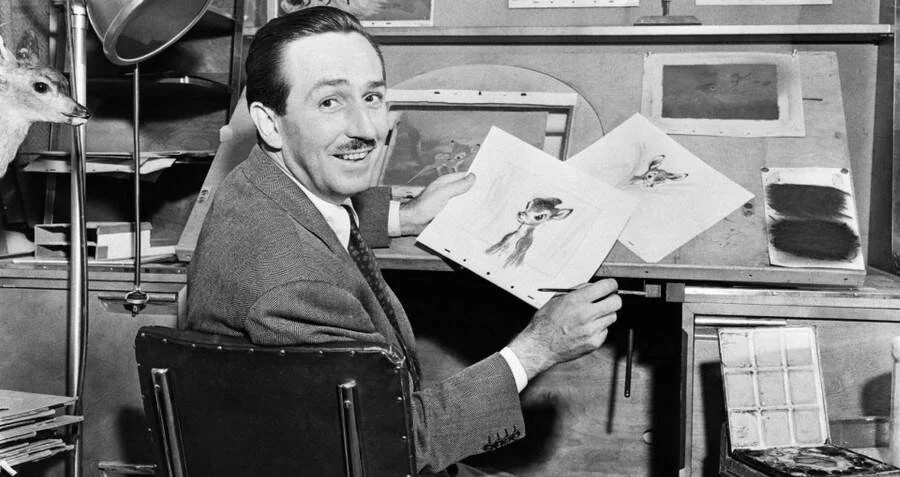 A man in a suit sits at a desk holding an animation sketch of a deer, surrounded by art materials and drawings, smiling at the camera in a studio setting.
