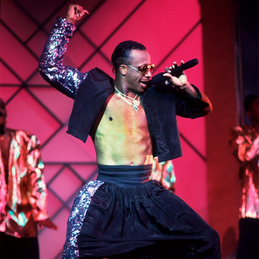 A performer wearing a sequined jacket, sunglasses, and loose black pants sings energetically into a microphone on a brightly lit stage with a pink and purple background.