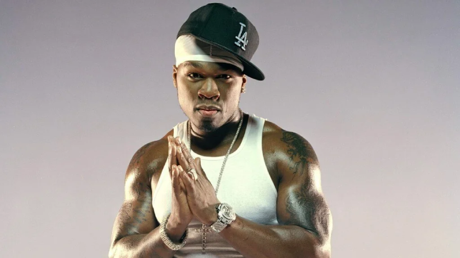A muscular man wearing a white tank top, large watch, and black LA baseball cap tilted to the side stands with his hands pressed together, looking confidently at the camera. He has visible tattoos and a white headband.