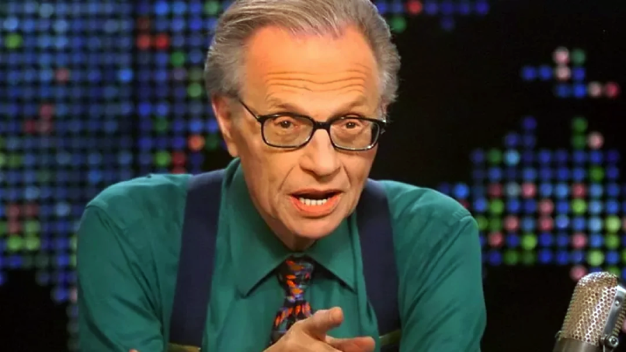 An older man with glasses, gray hair, and a green shirt with suspenders sits at a desk, speaking and gesturing with his hand. A colorful, pixelated background is behind him, and a microphone is visible in front.