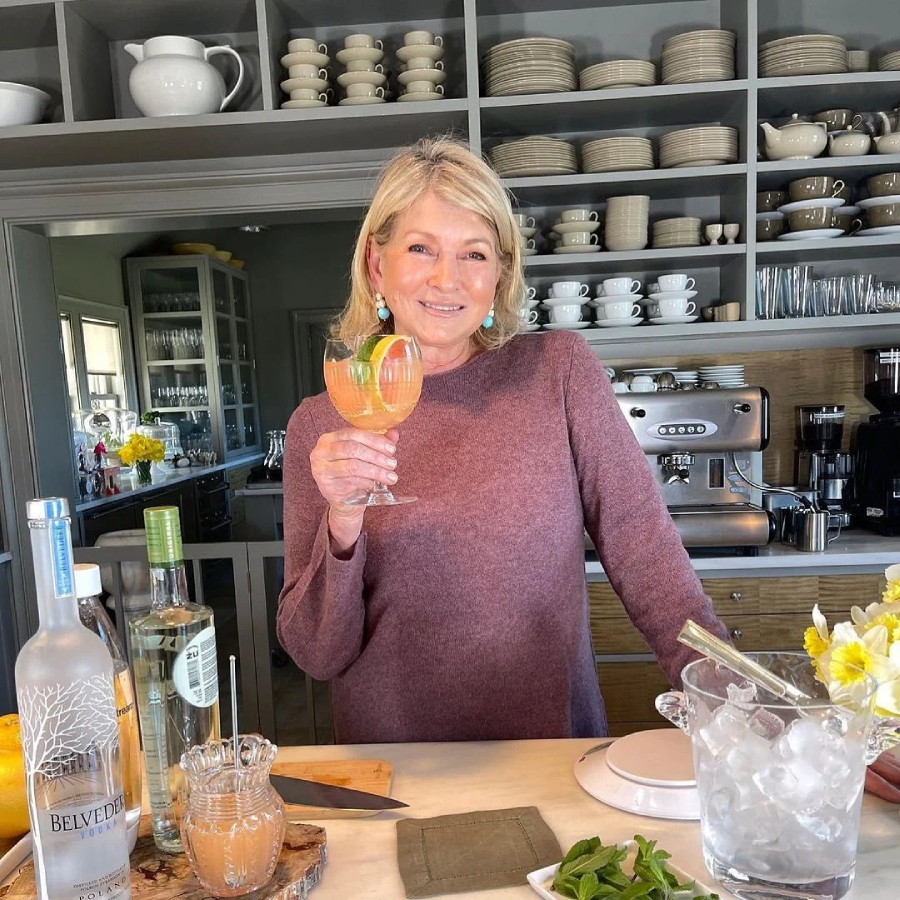 A woman in a mauve sweater stands in a modern kitchen, smiling and holding a pink cocktail. In front of her are bottles, a glass of ice, fresh mint, and a cutting board. Shelves of organized dishes fill the background.