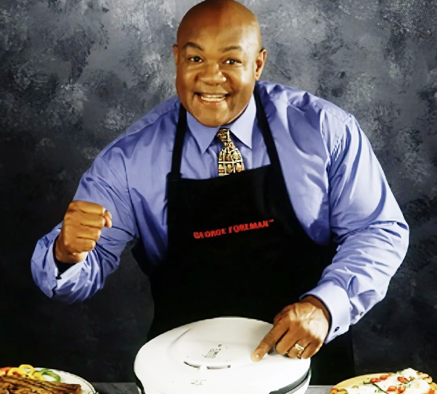 A smiling man in a blue shirt and patterned tie wears a black "George Foreman" apron, holding and pointing at a white countertop grill, with plates of food on either side against a dark background.