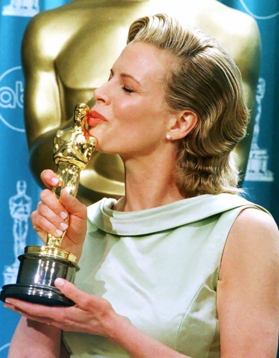 A woman in a light green dress kisses a gold Oscar statuette while standing in front of a blue backdrop with Oscar logos.