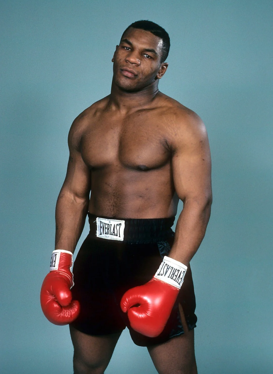 A muscular male boxer stands against a plain background, wearing red boxing gloves, black Everlast shorts, and no shirt. He looks directly at the camera with a serious expression.