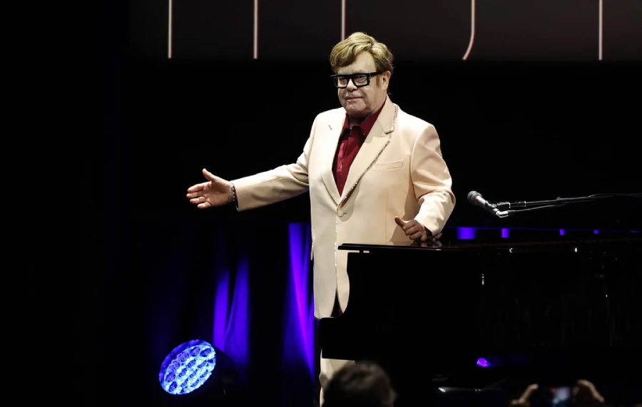 A person in a pale suit and red shirt stands on stage with arms open, next to a grand piano, under stage lights with a dark background.