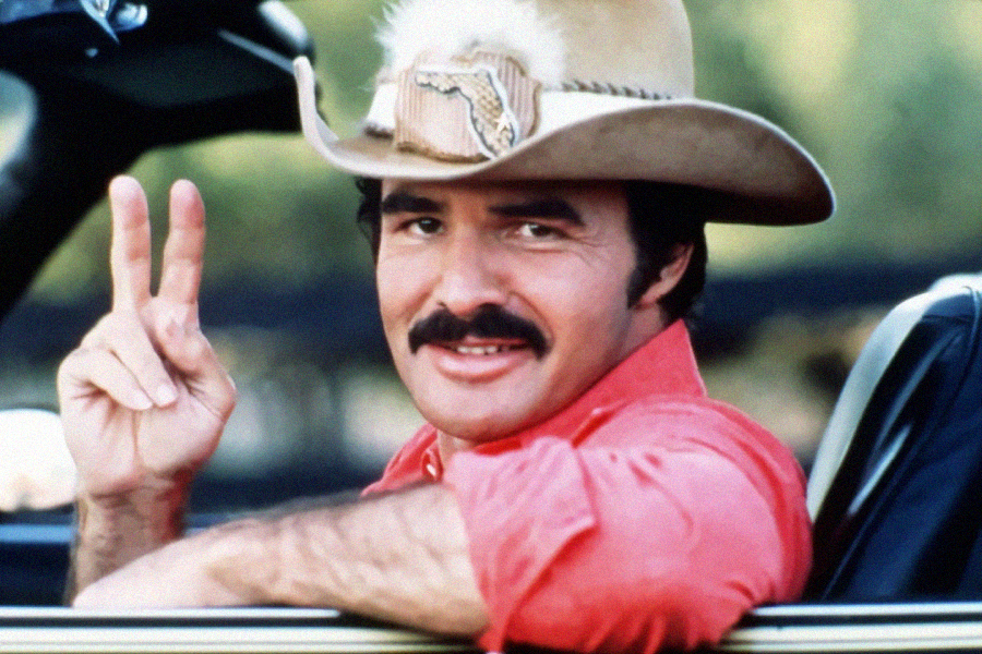 A man with a mustache wearing a tan cowboy hat and red shirt sits in a car, smiling and making a peace sign with his right hand.