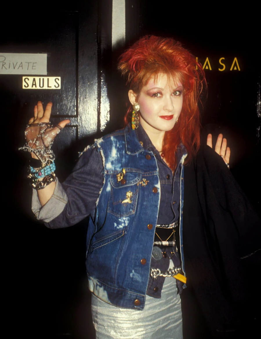 A woman with teased red hair, bold makeup, and colorful earrings poses with her hands up. She wears a sleeveless denim vest with pins, a studded glove, and layered bracelets, standing in front of a door labeled "Private Sauls.