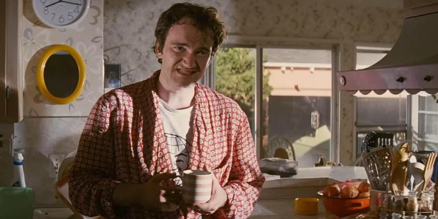 A man in a patterned red robe holds a striped mug while standing in a sunlit kitchen, with a window and clock visible in the background.