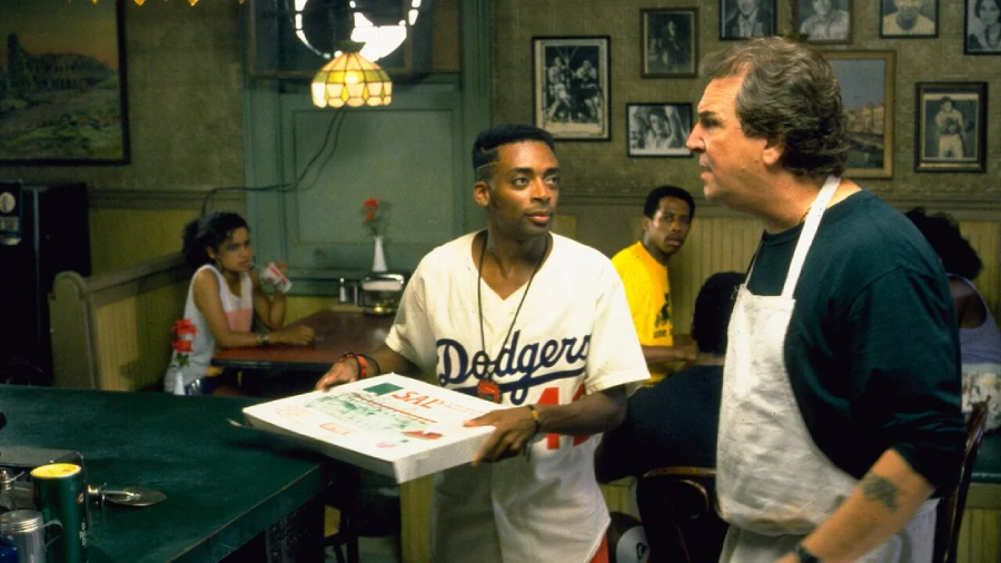 A man in a Dodgers jersey holds a pizza box and talks to a man in an apron behind a counter in a busy pizzeria, with customers sitting at tables in the background.