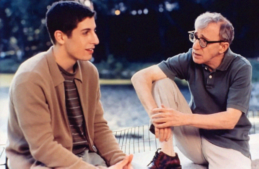 Two men sit outdoors having a conversation; one is young, wearing a brown jacket and striped shirt, while the older man in glasses and casual clothes listens intently with one knee raised. Trees and blurred scenery are in the background.