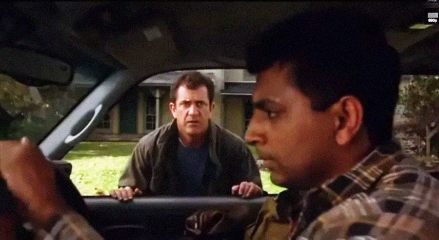 A man sits in a car, gripping the steering wheel, while another man stands outside the driver’s window, leaning on the door and looking at him with concern. A house and greenery are visible in the background.