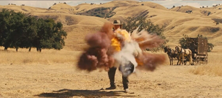 A person in a hat stands in a grassy field as a colorful explosion erupts around them. In the background, there are yellow hills, trees, and a horse-drawn wagon.