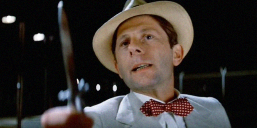 A man wearing a light-colored suit, red polka dot bow tie, and white hat points a knife forward, his expression serious, with a dark background behind him.