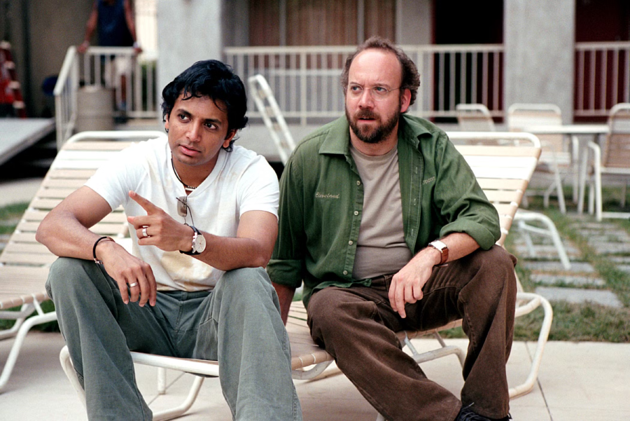 Two men sit on white lounge chairs outdoors; one wears a white t-shirt and light pants, pointing ahead, while the other wears glasses, a green shirt, and brown pants, looking in the same direction.