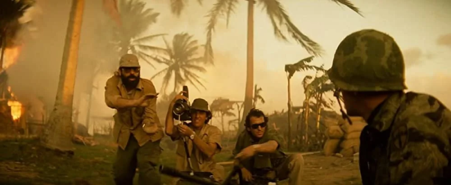 Three men, including a cameraman, film a soldier in a tropical war zone with palm trees, smoke, and fire in the background. The scene appears tense and chaotic.