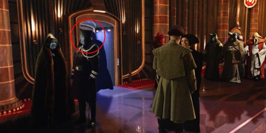 A group of people in elaborate costumes stand in a grand, ornately decorated hall; two figures in dark robes enter through a doorway on the left, one circled in red.