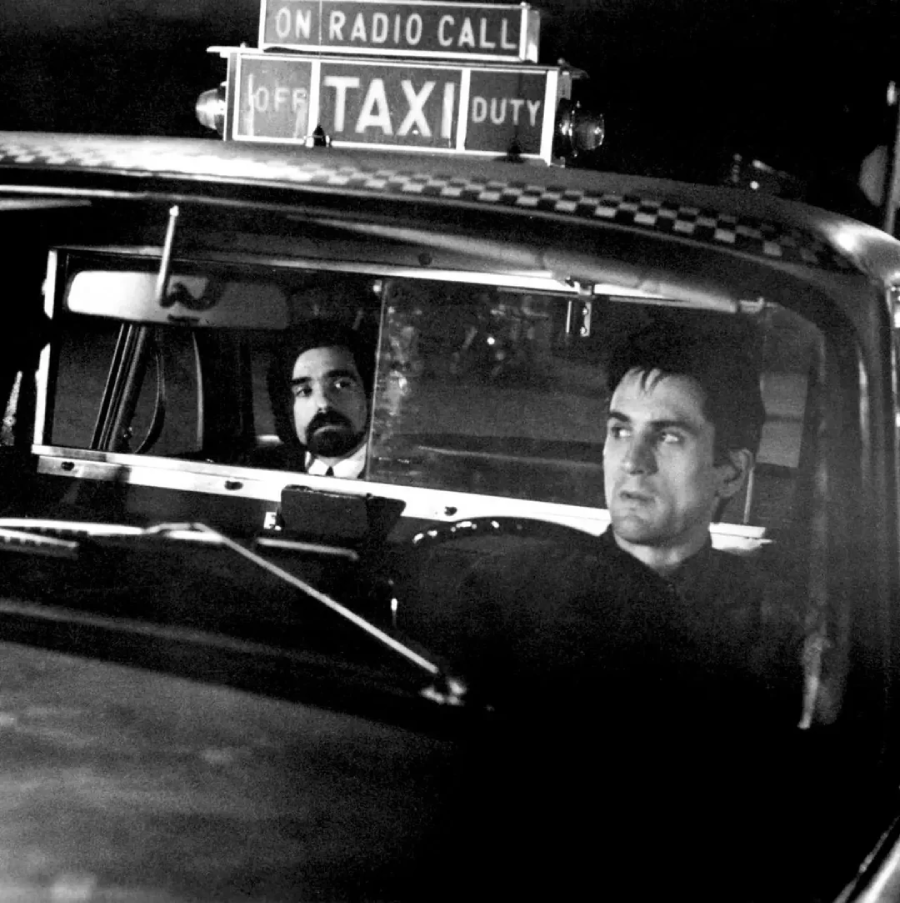 Black-and-white photo of two men in a taxi; one is driving and looking ahead, while the other sits in the back seat looking forward. A "TAXI" sign is visible on the roof of the car.