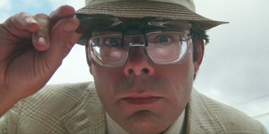 A man wearing large, thick glasses and a hat looks closely into the camera, lifting his glasses with one hand. He has a serious expression and is dressed in a light-colored jacket. The sky is visible in the background.
