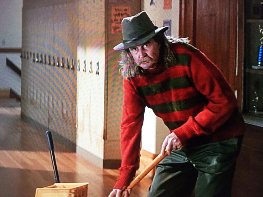 A man in a striped red and green sweater, olive pants, and a fedora mops the floor in a school hallway lined with lockers. He has long hair and a mustache, and looks up while cleaning.