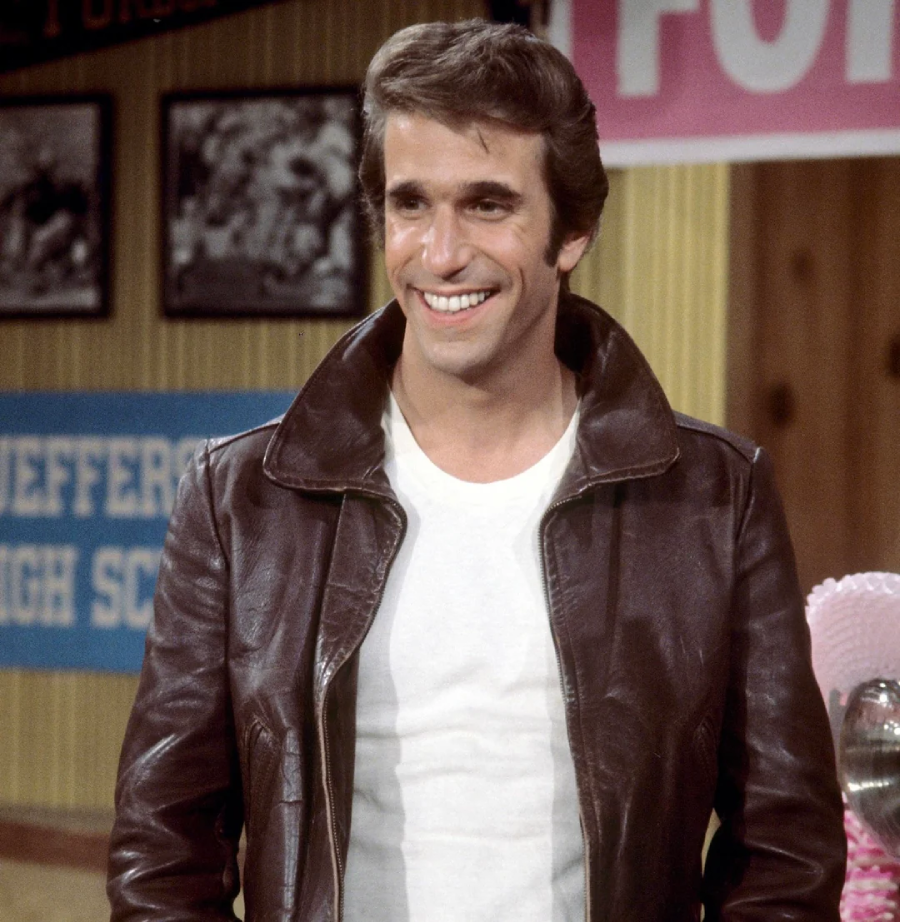A man with brown hair, wearing a brown leather jacket over a white t-shirt, smiles while standing indoors in front of a wall with framed photos and school banners.