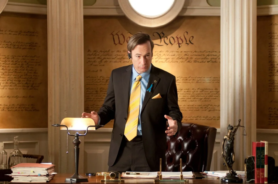 A man in a suit stands behind a cluttered desk in an office with columns and text from the U.S. Constitution on the wall. He has a serious expression and wears a blue ribbon on his lapel.