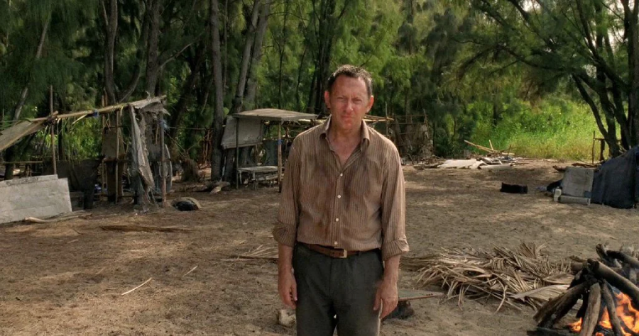 A man in a wrinkled, dirty shirt stands on sandy ground in front of makeshift huts and dense trees, suggesting a survival or deserted island scenario.