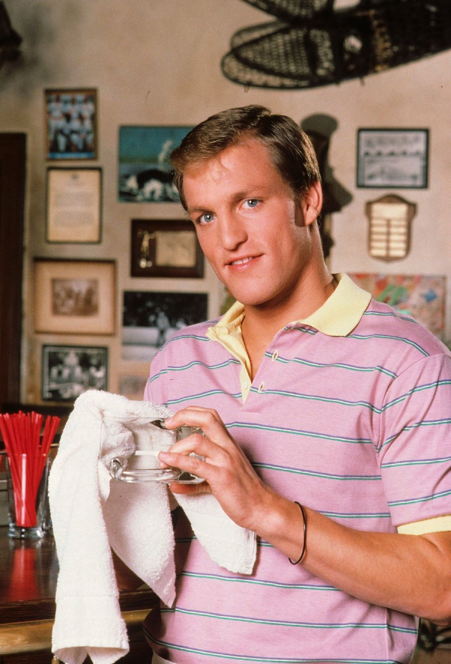 A smiling man in a pink striped polo shirt wipes a glass with a white towel while standing behind a bar, with framed photos and decorations on the wall in the background.