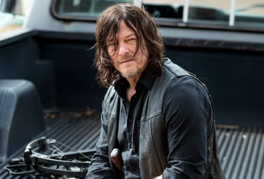 A man with long, messy brown hair and a scruffy beard sits in the bed of a pickup truck, wearing a black shirt and vest, holding a crossbow.