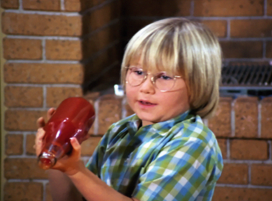 A young child with blonde hair and glasses holds a red ketchup bottle upside down. The child is wearing a green and blue plaid shirt and stands in front of a brick wall.