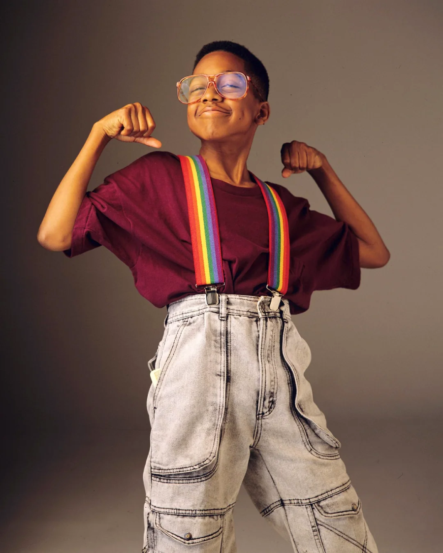 A smiling child wearing large glasses, a maroon shirt, rainbow suspenders, and high-waisted acid wash jeans poses confidently with arms flexed and fists up, standing against a plain background.