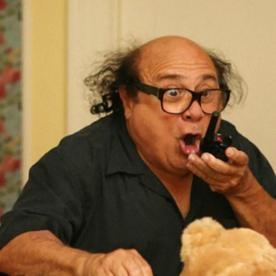 A surprised man with glasses and thinning hair holds a walkie-talkie close to his mouth, wearing a dark shirt. His mouth is open in shock and his eyes are wide. A blurry teddy bear is visible in the foreground.