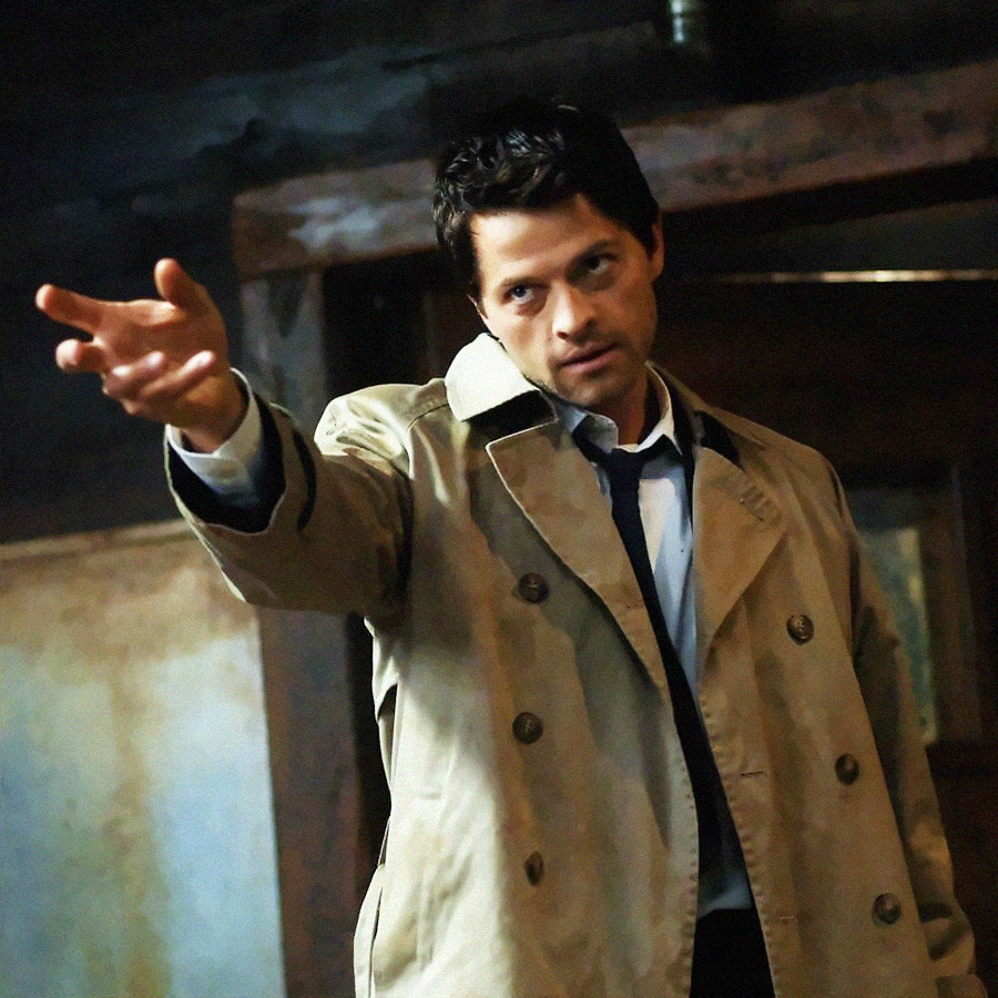 A man in a beige trench coat and blue shirt extends his arm, pointing with intensity. He has short dark hair and a serious expression, standing indoors in a dimly lit room.