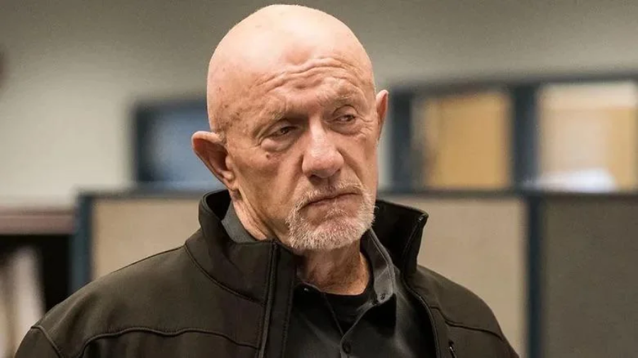A bald man with a serious expression, wearing a black jacket, stands indoors in front of blurred office cubicles.