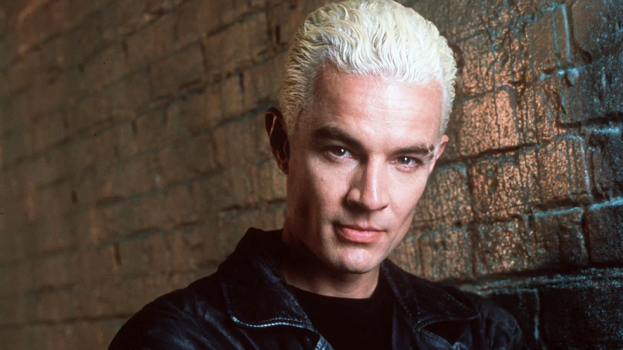 A man with slicked-back platinum blond hair and a black leather jacket stands against a brick wall, looking directly at the camera with a slight smirk.