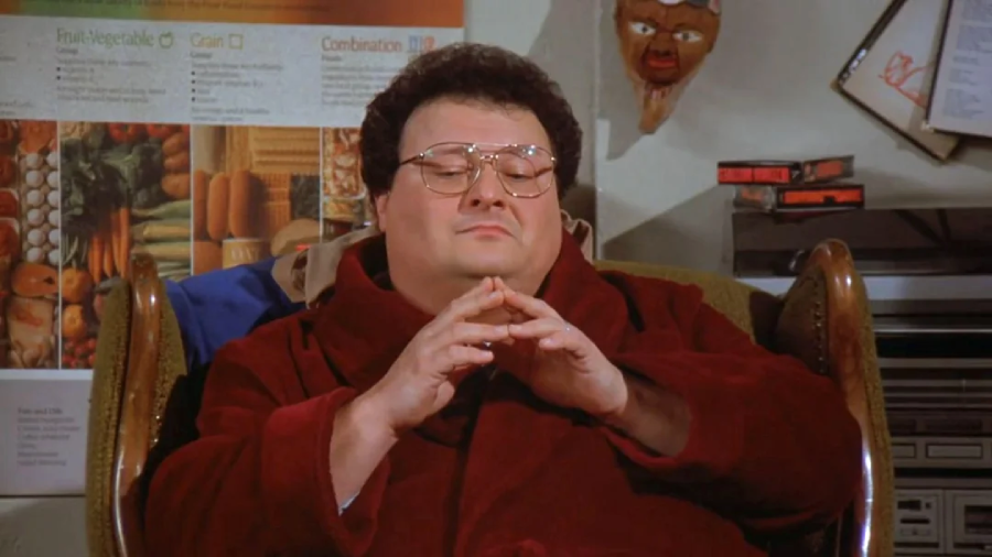 A man wearing glasses and a maroon robe sits with his fingers steepled, eyes closed, and an expression of satisfaction. Behind him are posters, a mask on the wall, and some audio equipment.