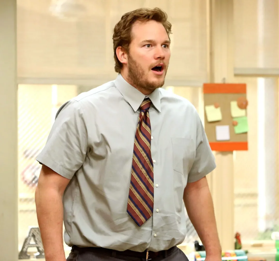 A surprised man with short brown hair and a beard, dressed in a gray dress shirt and striped tie, stands indoors with his mouth open, looking off to the side. Office supplies and notes are visible in the background.