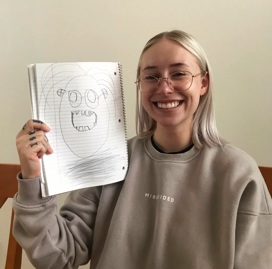 A smiling person with glasses and light hair holds up a notebook displaying a childlike drawing of a monster with big eyes and sharp teeth. They are wearing a beige sweatshirt and sitting indoors.