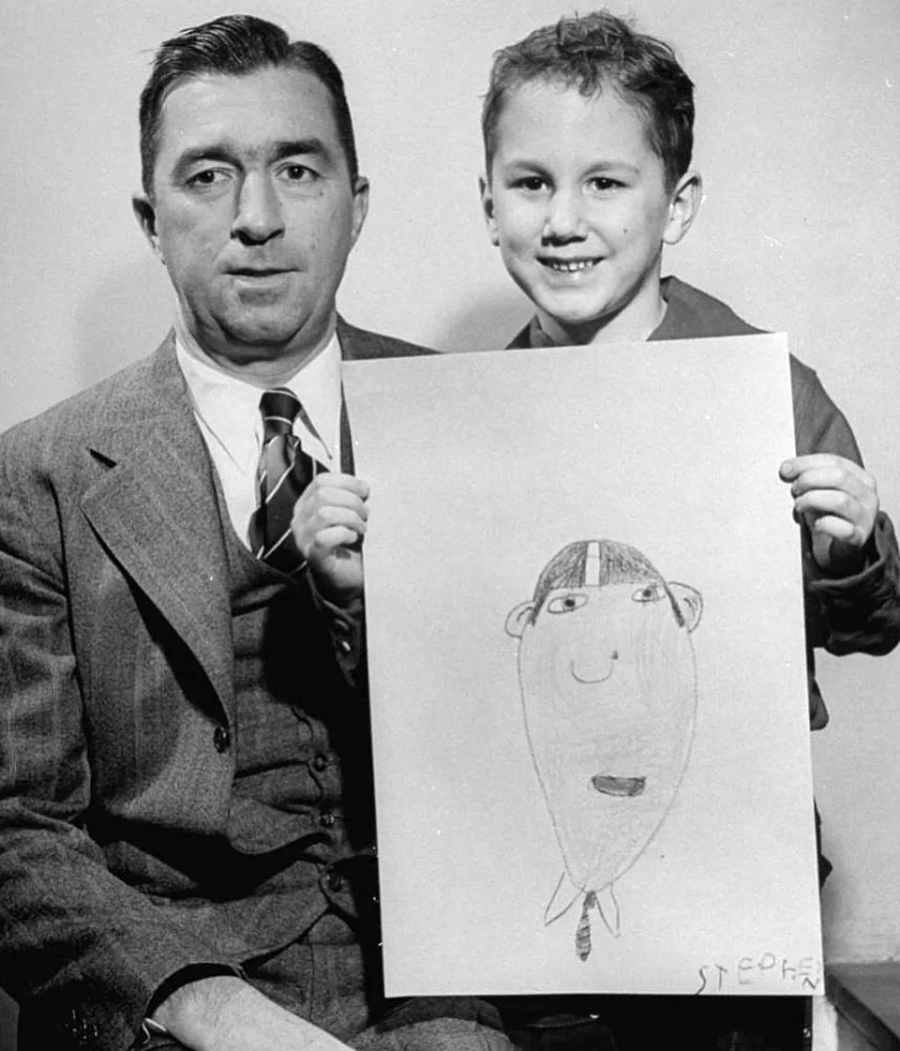 A man in a suit sits next to a smiling boy who is holding up a large sheet of paper with a simple, child’s drawing of a person wearing a cap.
