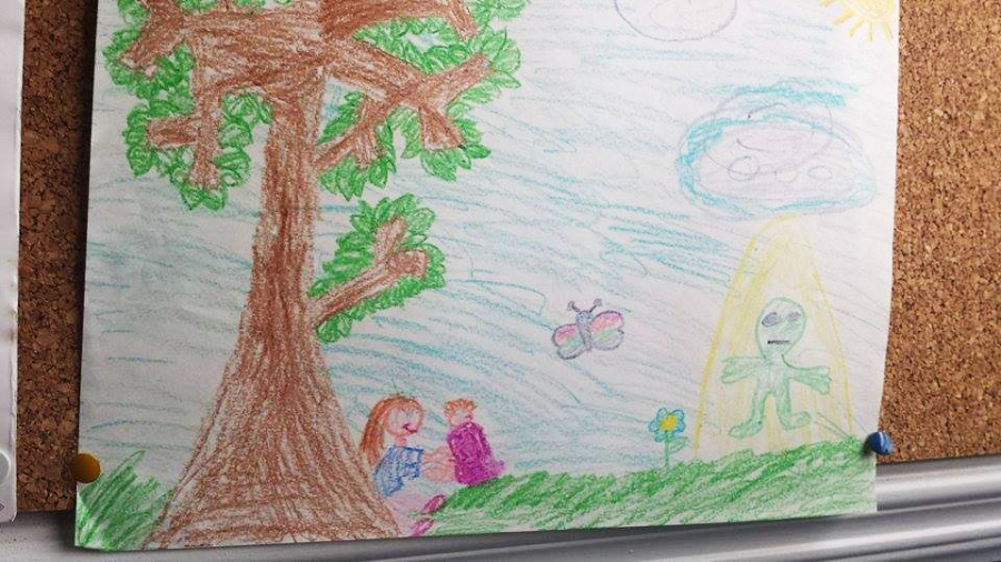 A child’s drawing shows two people sitting under a large tree, with a butterfly, a flower, and an alien figure standing in a yellow beam of light under a blue sky with clouds and a sun.