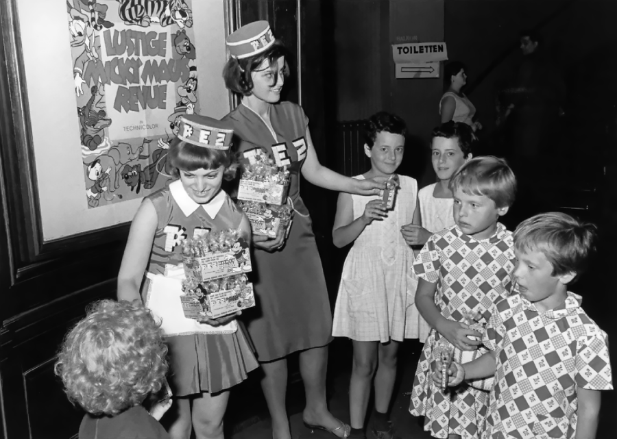 A woman and a girl wearing "FEZ" hats hand out snacks to a group of smiling children in dresses, standing indoors near a poster and a "Toiletten" restroom sign. The scene appears vintage, possibly from the 1960s.