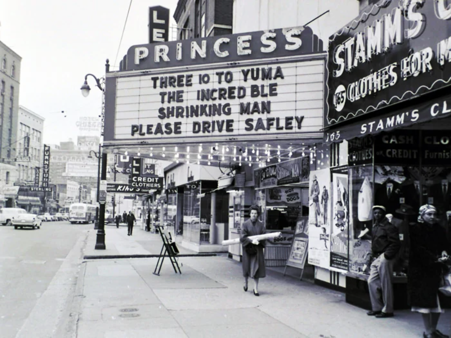 Black and white photo of a city street with the Princess Theater marquee displaying movie titles: "Three 10 to Yuma," "The Incredible Shrinking Man," and "Please Drive Safely." Pedestrians walk on the sidewalk.